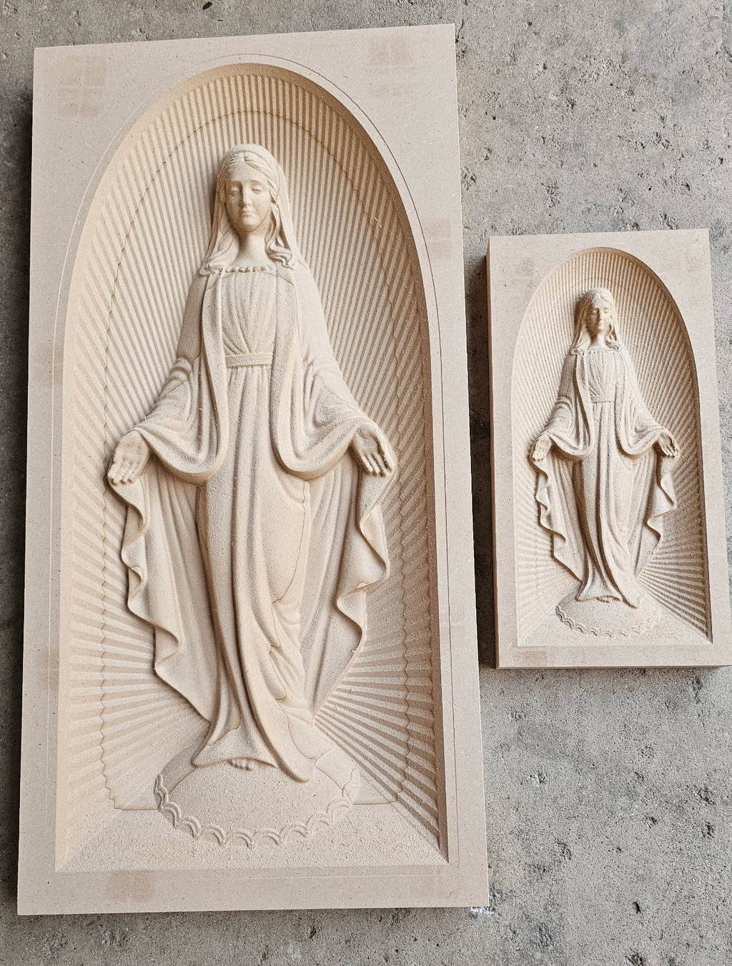 Two rectangular stone wall plaques featuring carved religious statues of the Virgin Mary in arched recesses, displayed on a textured gray concrete surface.
