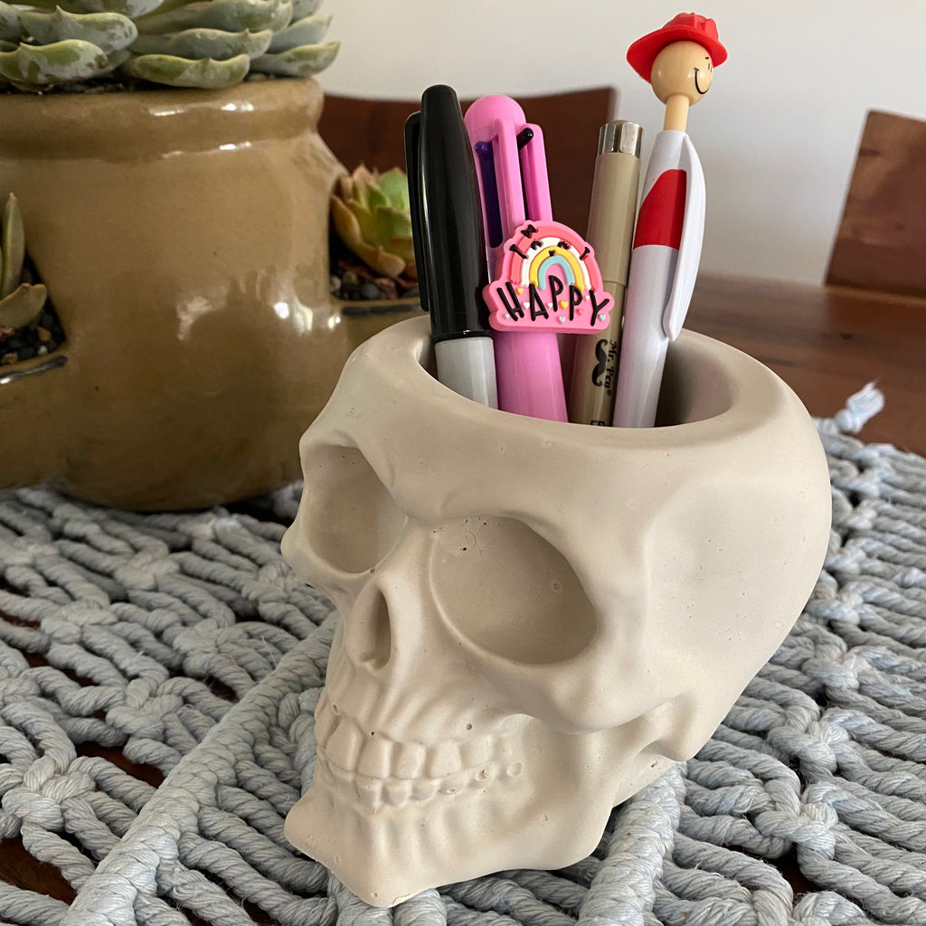 Skull-shaped pen holder with pens and a sticker on a textured surface