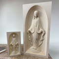 Two beige stone Virgin Mary wall plaques featuring carved female saint or Virgin figures in arched niches, displayed on a tabletop against a plain light background.