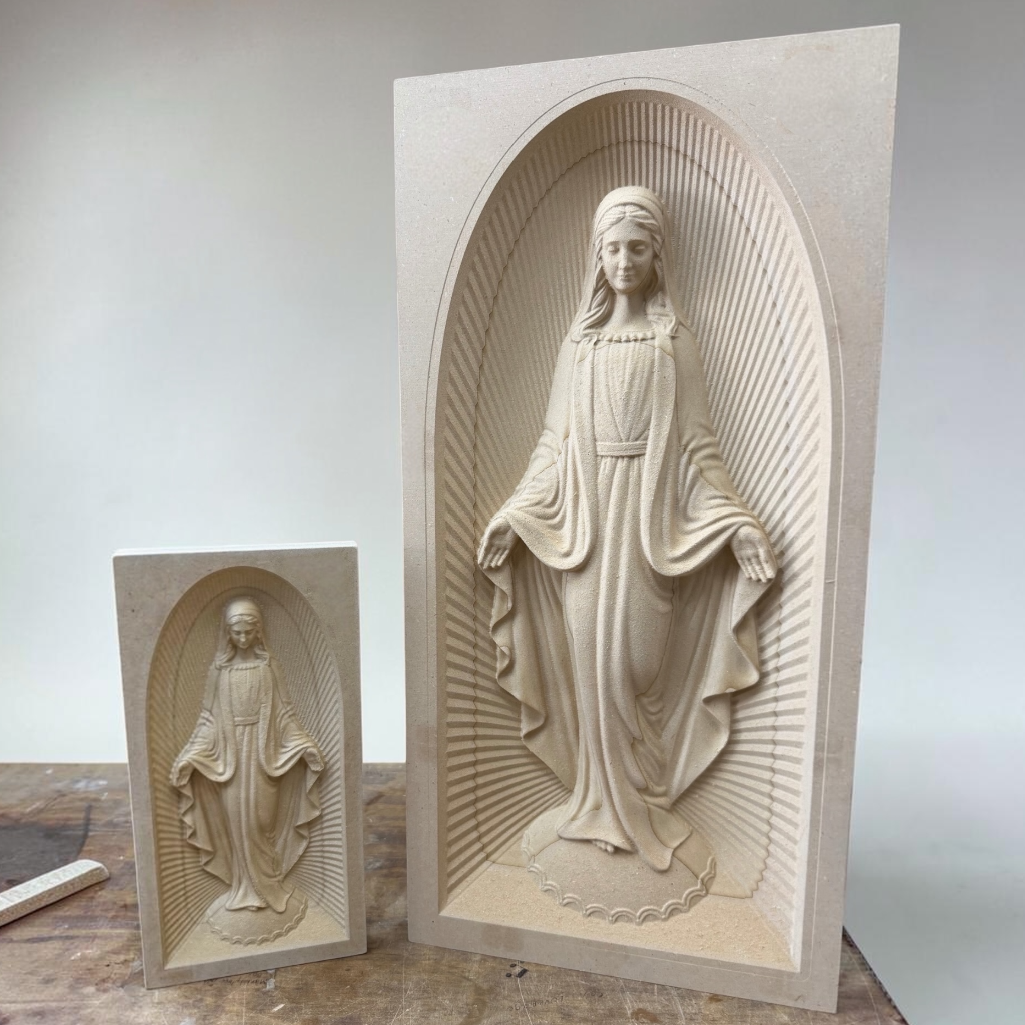Two beige stone Virgin Mary wall plaques featuring carved female saint or Virgin figures in arched niches, displayed on a tabletop against a plain light background.
