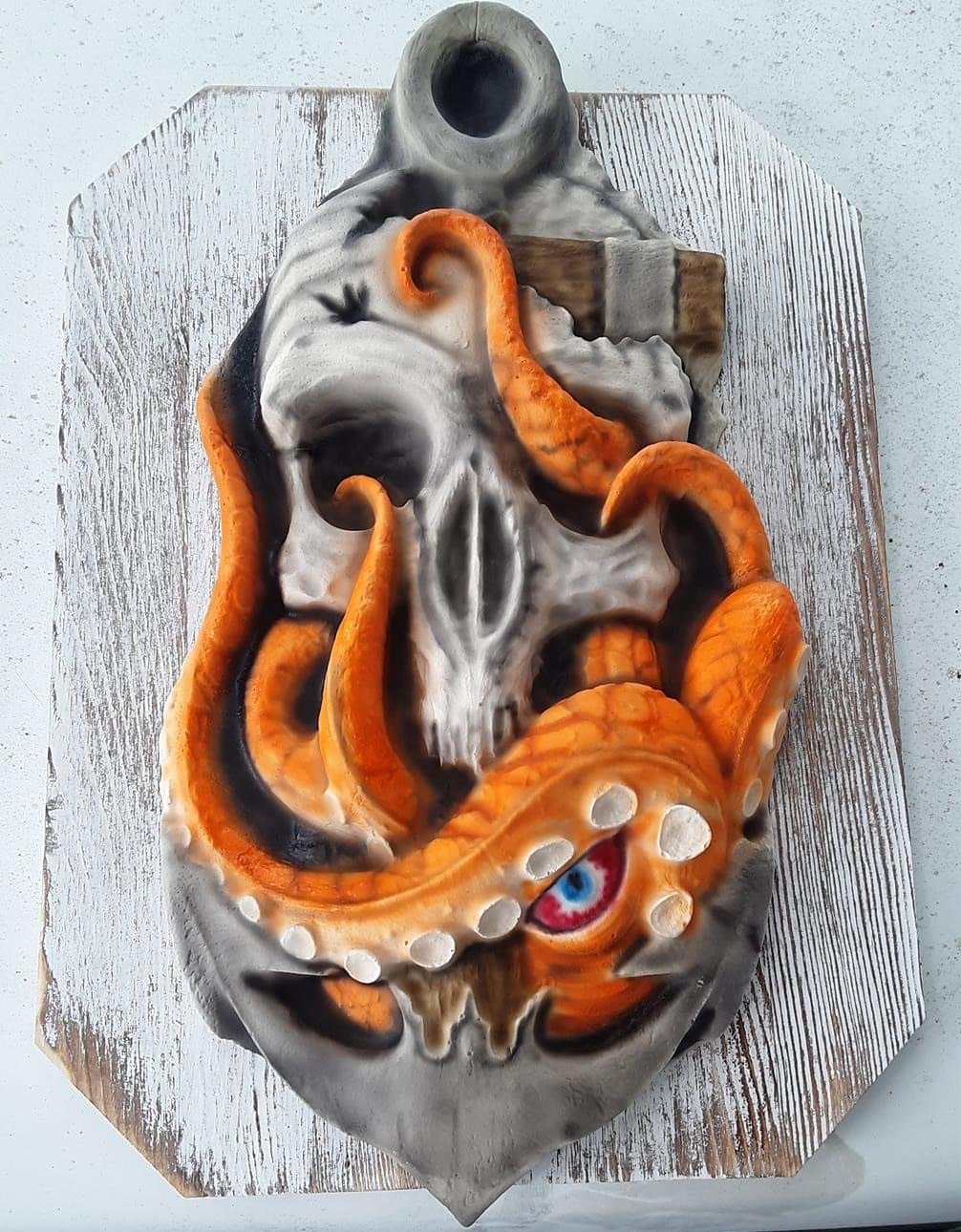 A mold of a skull with an anchor blasted into it, and an octopus with orange tentacles wrapped around the anchor and skull.