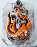 A mold of a skull with an anchor blasted into it, and an octopus with orange tentacles wrapped around the anchor and skull.